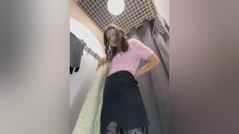 Media: Video of a woman in a pink sweater, black skirt, and white stockings, standing in a bathroom with a beige carpet, grey curtains, and a circular light fixture.