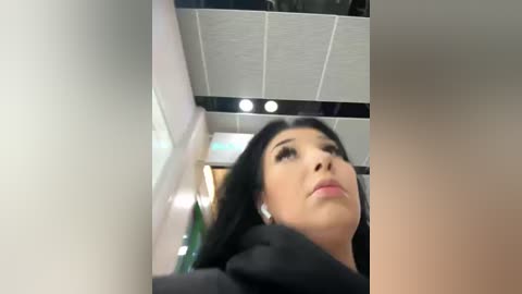 Media: Video of a woman with long black hair and light skin, wearing black clothes, looking upwards. Background features a modern, tiled ceiling with recessed lights and a blurred office setting.