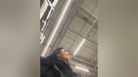 Media: Video of a young Asian woman with black hair, wearing a black jacket, standing in a dimly lit industrial warehouse with a high, corrugated metal ceiling and exposed pipes.