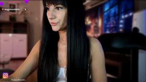 Media: A video of a young, slender woman with long, straight black hair and fair skin, wearing a white lace top. She stands indoors near a window with a city skyline view.