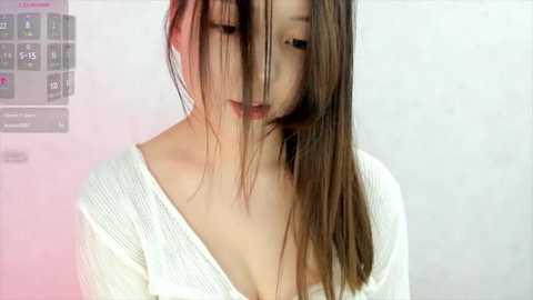 Media: A young Asian woman with long, straight brown hair partially covering her face, wearing a white ribbed sweater with a deep V-neck. The background is a blurred, light-colored wall.