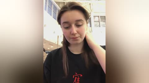 Media: Video of a young woman with long brown hair, wearing a black shirt with a red graphic, standing in a brightly lit indoor space with white walls and large windows.