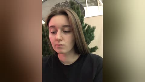 Media: Video of a young, light-skinned Caucasian woman with straight, medium-length brown hair, wearing a black shirt. Background features a green Christmas tree and beige walls with a circular window.