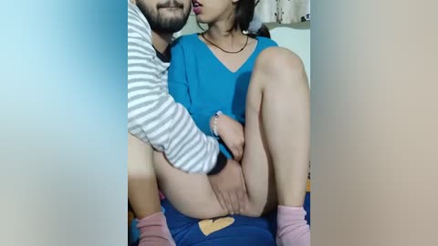 Media: A video shows an Asian woman in a blue top and pink socks, sitting on a man's lap, with her legs spread and his hand between her thighs.