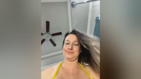 Media: A video of a plus-sized woman with long, wavy brown hair, wearing a yellow bikini top, standing in a bathroom with a ceiling fan and shower.