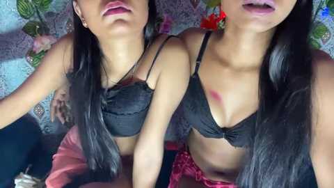 Media: Video of two young women with long black hair, wearing black bras and red pants, lying on a patterned surface.