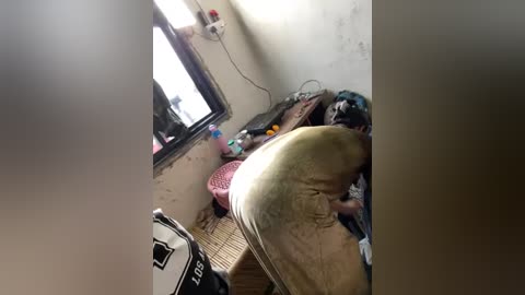 Media: Video of a messy, cramped room with a man in a light-colored shirt and tan pants. The room has a window, a table, and various items scattered around.