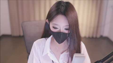 Media: A video of an East Asian woman with long, straight black hair, wearing a white blouse, black face mask, and black headphones, sitting indoors, focusing on her phone.