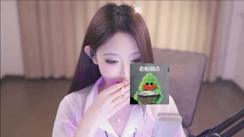 Media: Video of an East Asian woman with long brown hair, wearing a white blouse, examining a smartphone with a green duck emoji on the screen. She sits indoors, with beige curtains in the background.