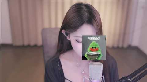 Media: Video of a young Asian woman with long brown hair, wearing a black top, sitting at a microphone, blurred background with beige curtains.