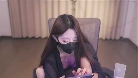 Media: A video of a young Asian woman with long black hair, wearing a black mask, sitting at a desk in a room with beige curtains and a wooden floor.