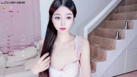 Media: Video of a young Asian woman with pale skin, long black hair, and pink lips, wearing a light pink lace bra, standing next to a white staircase in a modern, minimalist room.