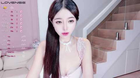 Video of a slim East Asian woman with long black hair, wearing a white lace lingerie set with a choker, standing in a modern, pink-themed room with a white staircase.