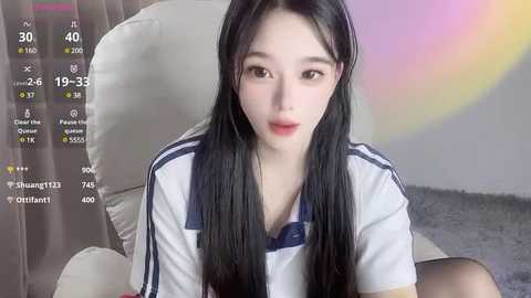 Media: Video of an East Asian woman with long black hair, fair skin, wearing a white and blue striped shirt, sitting on a beige couch, with a virtual overlay displaying her stats and a rainbow effect.