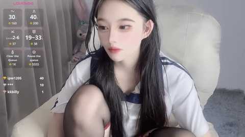 Media: Video of a young East Asian woman with long black hair, wearing a white sailor outfit, sitting on a white leather chair. She has a pale complexion and a contemplative expression. The background is a light-colored room with a window and a carpet.