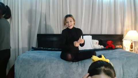 Media: Video of a blonde woman in a black dress and red hairband, sitting on a blue bed with a black headboard, talking to someone off-camera, surrounded by stuffed toys and a lamp.