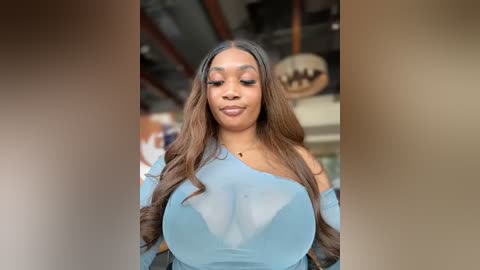 Media: Video of a confident Black woman with long, wavy hair, wearing a sheer, light blue top that highlights her large breasts. She's indoors, with a blurred background of modern, industrial decor.