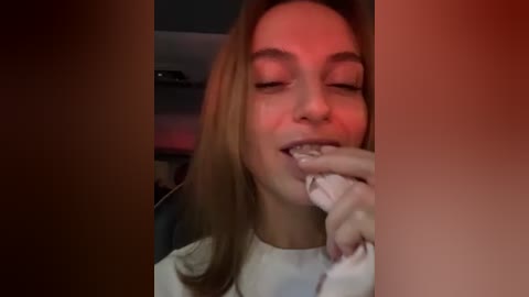 Media: Video of a young woman with light skin, straight brown hair, wearing a white shirt, biting her fingernail in a dimly lit room, red lighting, blurry background.