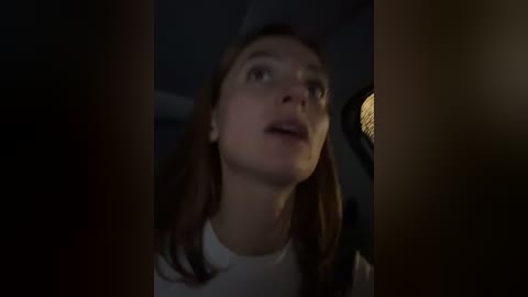 Media: A video of a Caucasian woman with long brown hair, wearing a white top, looking up in surprise with a dimly lit room in the background.