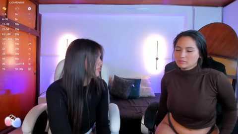 Media: Video of two women in a dimly lit room with a digital clock displaying 10:10. One wears a black turtleneck, the other a brown turtleneck, both with long dark hair.