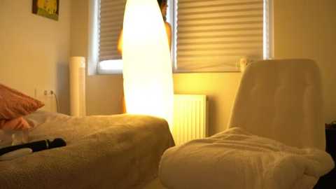 Media: A video of a cozy bedroom with two beds, each covered in white blankets, and a large, glowing white lamp casting a warm, soft light on the white walls and window blinds.