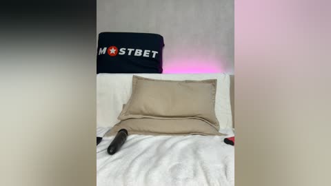 Media: A video of a bed with beige pillows and a white duvet, featuring a dark pillow with \"MSTBET\" in white and red letters. The background is a gradient of black, white, and pink.