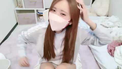 Media: Video of a young East Asian woman with long brown hair, wearing a white face mask, sitting on a bed, holding her hair, in a minimalist, white-walled room.