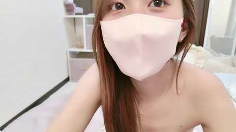 Media: Video of an Asian woman with long brown hair, wearing a pink face mask, standing in a bedroom with white walls and a bed in the background.