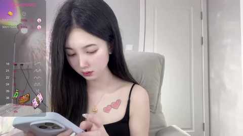 Media: Video of a young Asian woman with long black hair, fair skin, and a slim physique, wearing a black tank top, sitting on a beige chair, intently using a smartphone with a white case.