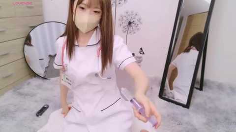 Media: A young Asian woman with light skin and long brown hair, wearing a white nurse outfit, mask, and holding a spray can, kneeling in a minimalist room with white walls, a mirror, and a dresser.