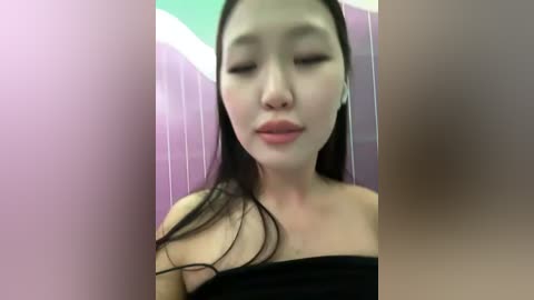 Media: Video of an Asian woman with long black hair and pale skin, wearing a black strapless top, looking serene with closed eyes. Background features a purple-striped wall and a green ceiling.