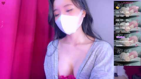 Media: A video of an Asian woman with long black hair, wearing a white mask, revealing cleavage in a low-cut, grey cardigan. Background includes a pink curtain and a pink room with a bed.