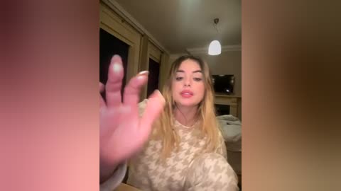 Media: Video of a young Caucasian woman with long blonde hair, wearing a beige floral sweater, holding her hand up in a bedroom with a bed, shelves, and a hanging light fixture.