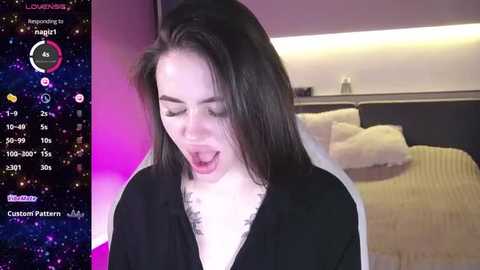 Media: Video of a woman with long, straight black hair, wearing a black dress, mouth open in an exaggerated expression of surprise or shock. Background shows a bedroom with a bed, nightstand, and a colorful digital display.