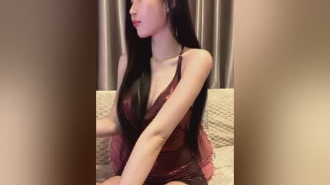 Media: Video of an East Asian woman with long, straight black hair, wearing a revealing, sheer burgundy lingerie dress with lace trim, sitting on a beige textured couch.