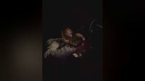 Media: A dimly lit video of a woman in a white jacket, with her face partially illuminated, driving at night. The background is dark, creating a dramatic, suspenseful atmosphere.