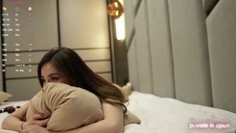 Media: Video of a young woman with long brown hair, lying on a bed with beige pillows, wearing a beige top, in a modern bedroom with a soft, warm ambiance.