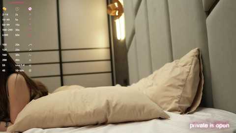 Media: Video of a woman lying face down on a beige bed with a gray padded headboard, partially covered by a cream-colored pillow. A modern, minimalist room with a sliding shoji screen and a gold pendant lamp in the background.