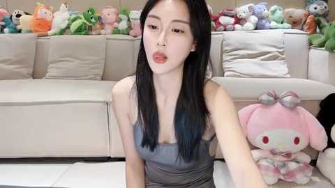 Media: Video of an East Asian woman with long black hair, light skin, and a petite frame, wearing a sleeveless striped top, seated in a plush, beige-furnished room with plush toys and stuffed animals in the background.