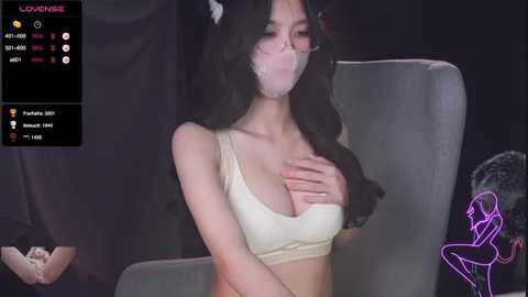 Media: A video captures an Asian woman with long black hair and pale skin, wearing a beige sports bra and tape over her mouth. She sits on a gray chair in a dimly lit room, with a neon pink bunny icon in the corner.