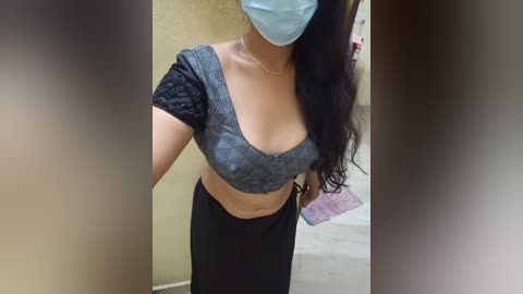 Media: Video of a woman with medium skin tone and long black hair, wearing a light blue face mask, a grey crop top with black lace sleeves, and black pants, standing in a beige-tiled room.