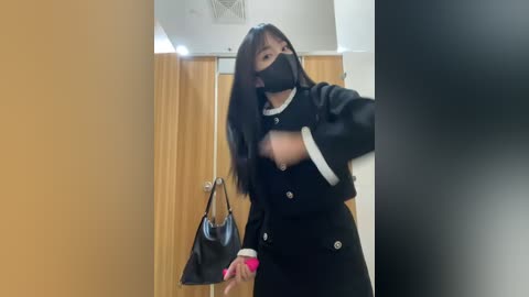 Media: Video of a woman with long black hair, wearing a black face mask, black jacket, white shirt, and holding a black handbag. She stands in a dimly lit, beige-tiled room with a wooden door and curtain.