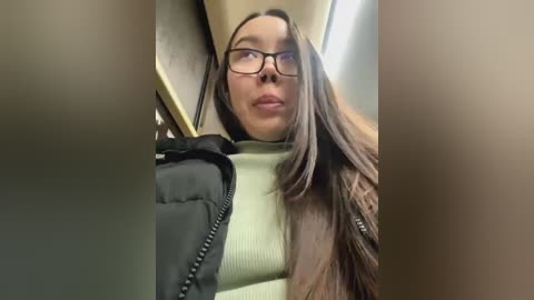 Media: Video of a young woman with long brown hair, wearing black-rimmed glasses and a beige sweater, sitting on a train. She appears to be looking upwards, slightly smiling. The background includes train seats and a window.