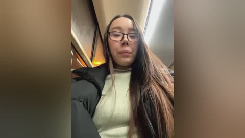 Media: Video of a young woman with long, straight brown hair, wearing black-framed glasses, a black jacket, and a cream turtleneck sweater. She's sitting in a bus, with beige walls and overhead lights in the background.