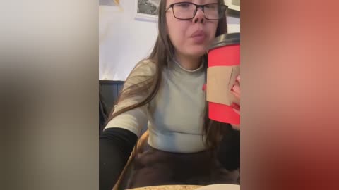 Media: Video of a young woman with long brown hair, wearing glasses, a light grey shirt, and black gloves, holding a red coffee cup, sitting indoors, blurry background.