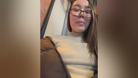 Media: Video of a young woman with long brown hair, wearing glasses and a beige sweater, looking relaxed in a car.