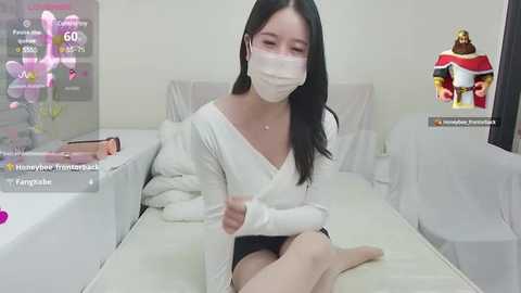 Media: Video of an East Asian woman with long black hair, wearing a white mask and off-shoulder top, sitting on a white bed in a minimalist room.