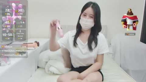 Media: A video of an East Asian woman with long black hair, wearing a white mask and T-shirt, sitting on a bed, holding a pink object. A digital overlay shows a character in a video game.