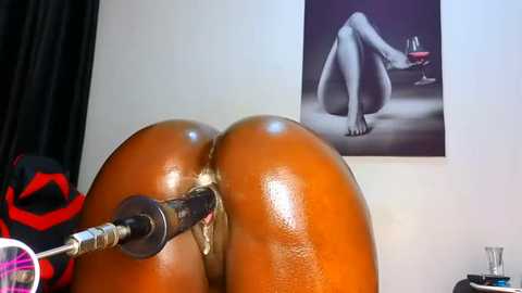 Media: Video of a dark-skinned person with a large, shiny, bare buttocks being penetrated by a metal butt plug, set against a white wall with a black-and-white wine glass painting.