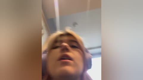 Media: A blurry video of a blonde woman with short hair, possibly in her 20s, standing inside a white-walled room with fluorescent lighting, making a silly face.